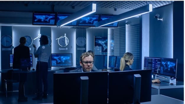 A group of cybersecurity professionals working in a high-tech security operations center. The room is filled with large screens displaying network security data and threat analysis reports.